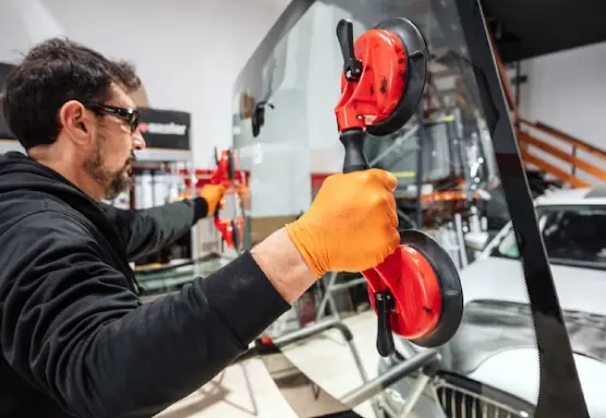 A person wearing black goggles and orange gloves is holding a car windshield with the help of two suction cups.